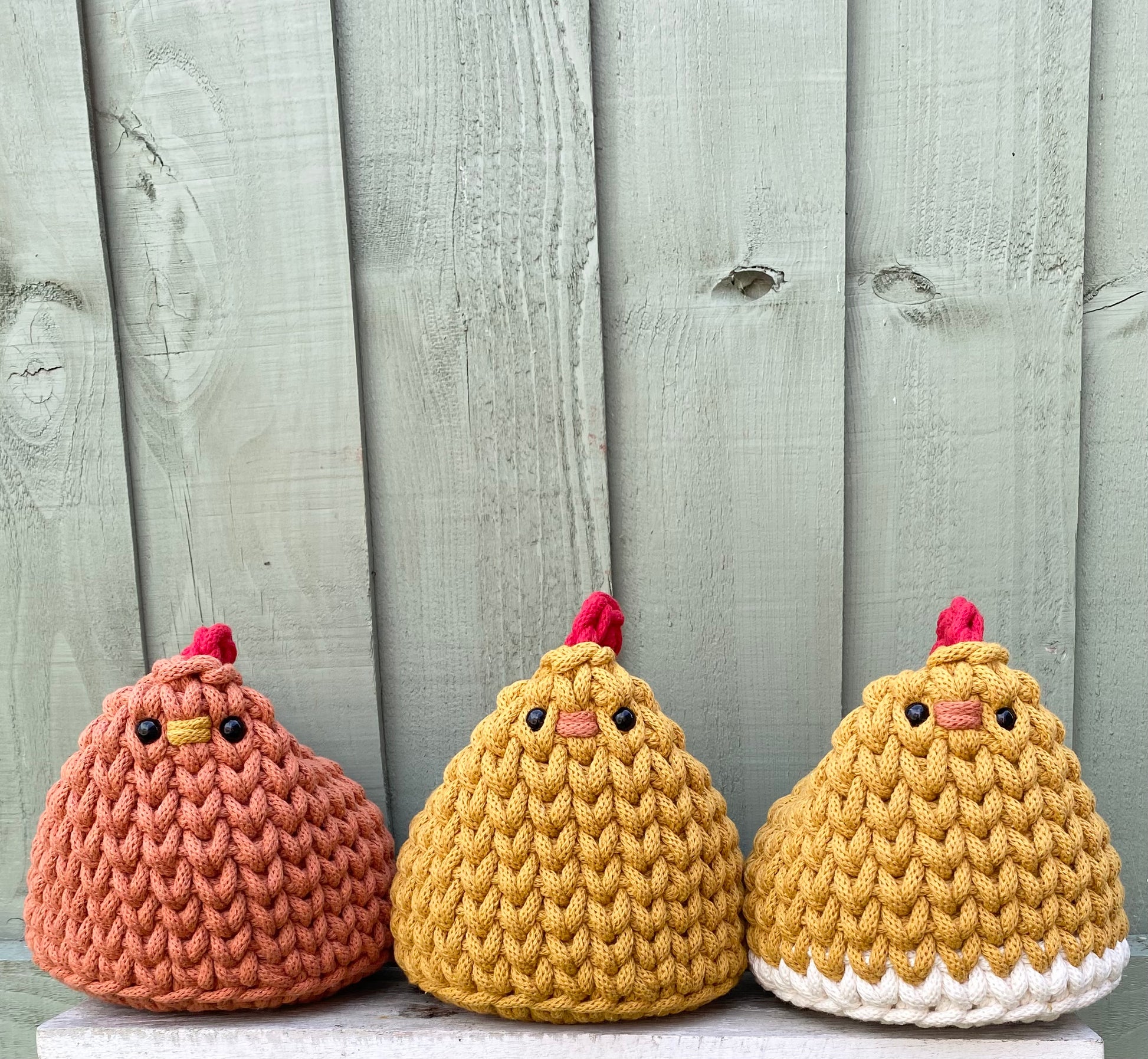 Three Henrietta the hen chicken doorstops in different colour's against a wooden background
